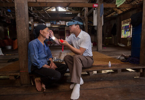 A Vietnamese women on the left has her eyelids inverted to check for sign of trachoma infection by a health worker wearing loupes and holding a flashlight. 